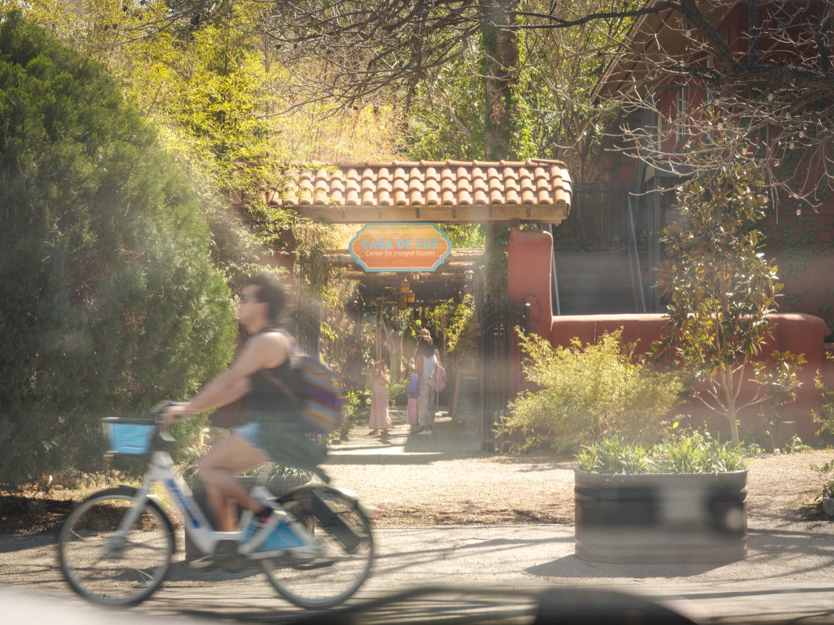 The tree-shaded entrance to Casa de Luz on Toomey Road in Austin, with a cyclist passing on the street